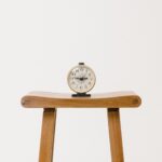 An analog clock with black numbers and hands sits on a wooden stool against a plain white background. The clock displays the time as 10:10.