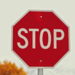 A red octagonal stop sign with "STOP" written in white capital letters. The background features a blurred tree with orange leaves.