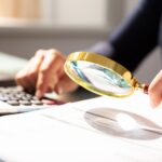 A person uses a magnifying glass to closely examine documents, with a calculator nearby on the desk. The scene suggests careful inspection and analysis of financial or business information.