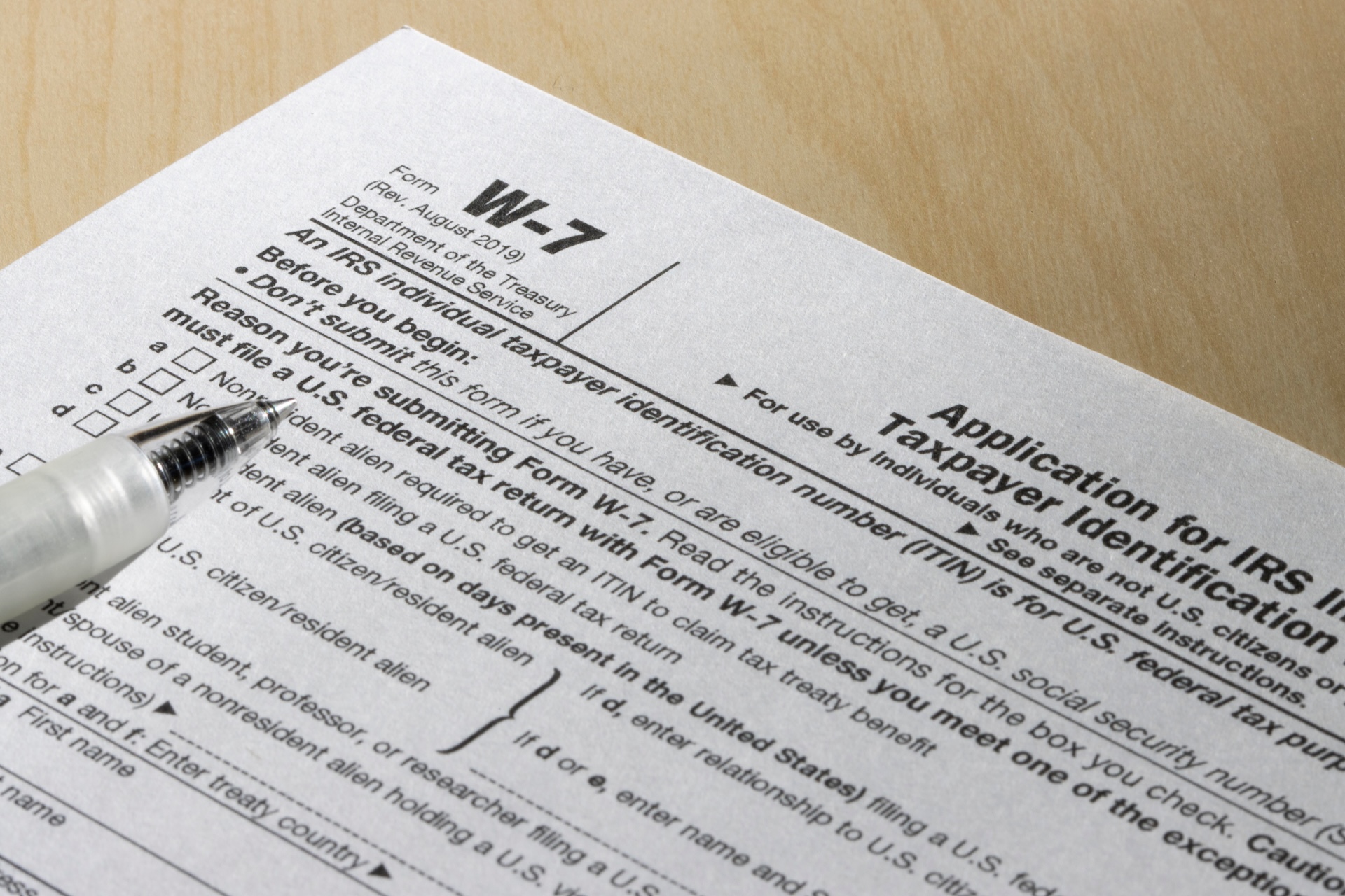 A close-up photo of an IRS Form W-7, "Application for IRS Individual Taxpayer Identification Number," resting on a wooden surface. A pen is positioned nearby, poised to fill out the form.