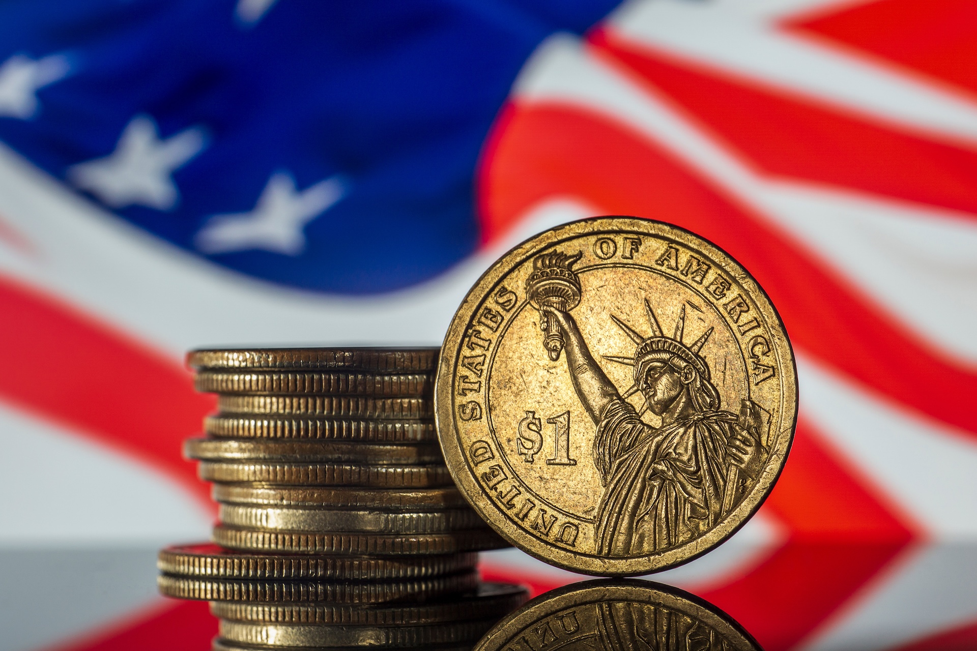 A stack of U.S. one dollar coins featuring the Statue of Liberty, with an American flag in the background. The coins are gold-colored and prominently display "$1" and "United States of America.