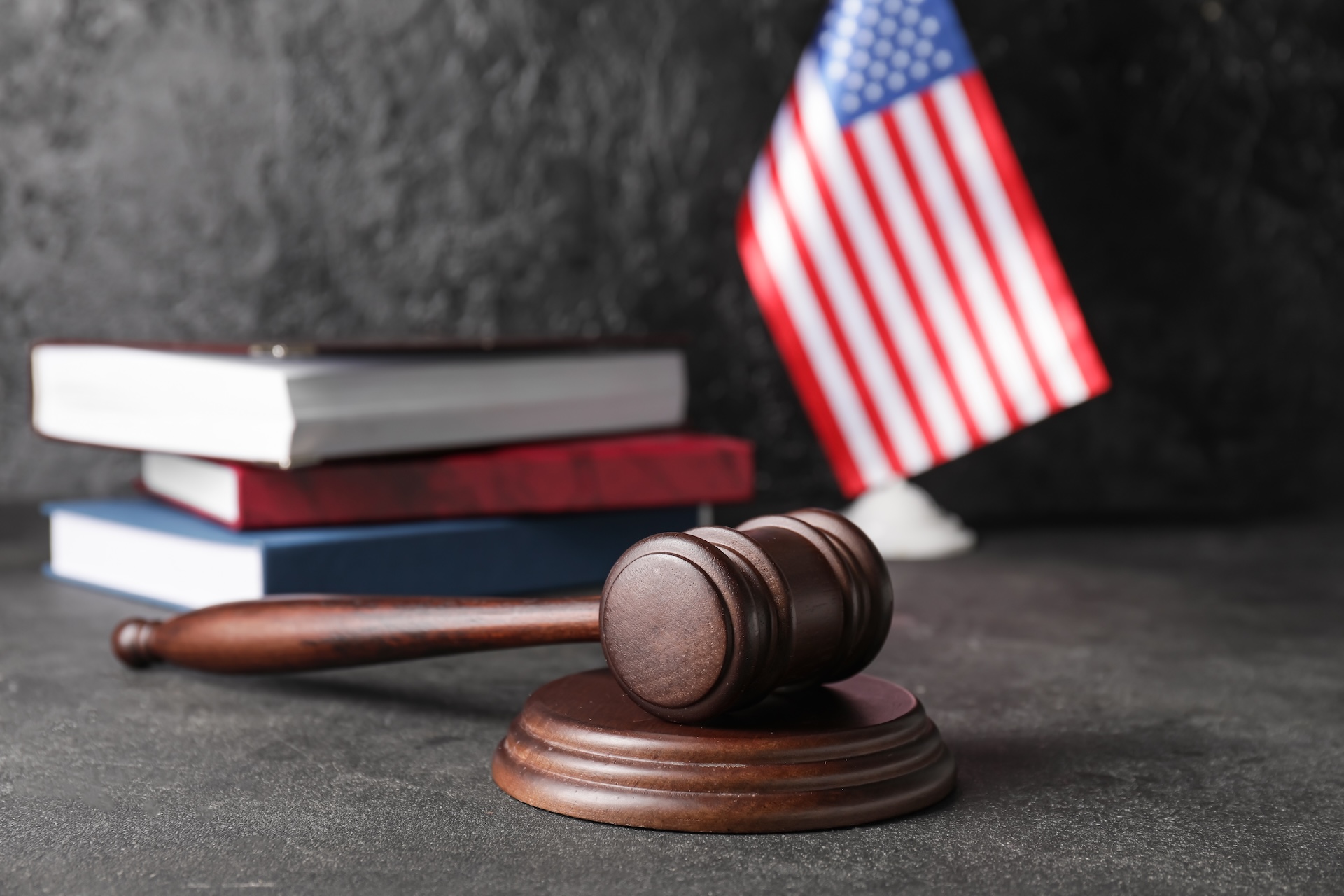 A wooden gavel rests on a sound block in the foreground. Behind it are two stacked books with red and blue covers. An American flag is visible in the background against a textured dark wall.