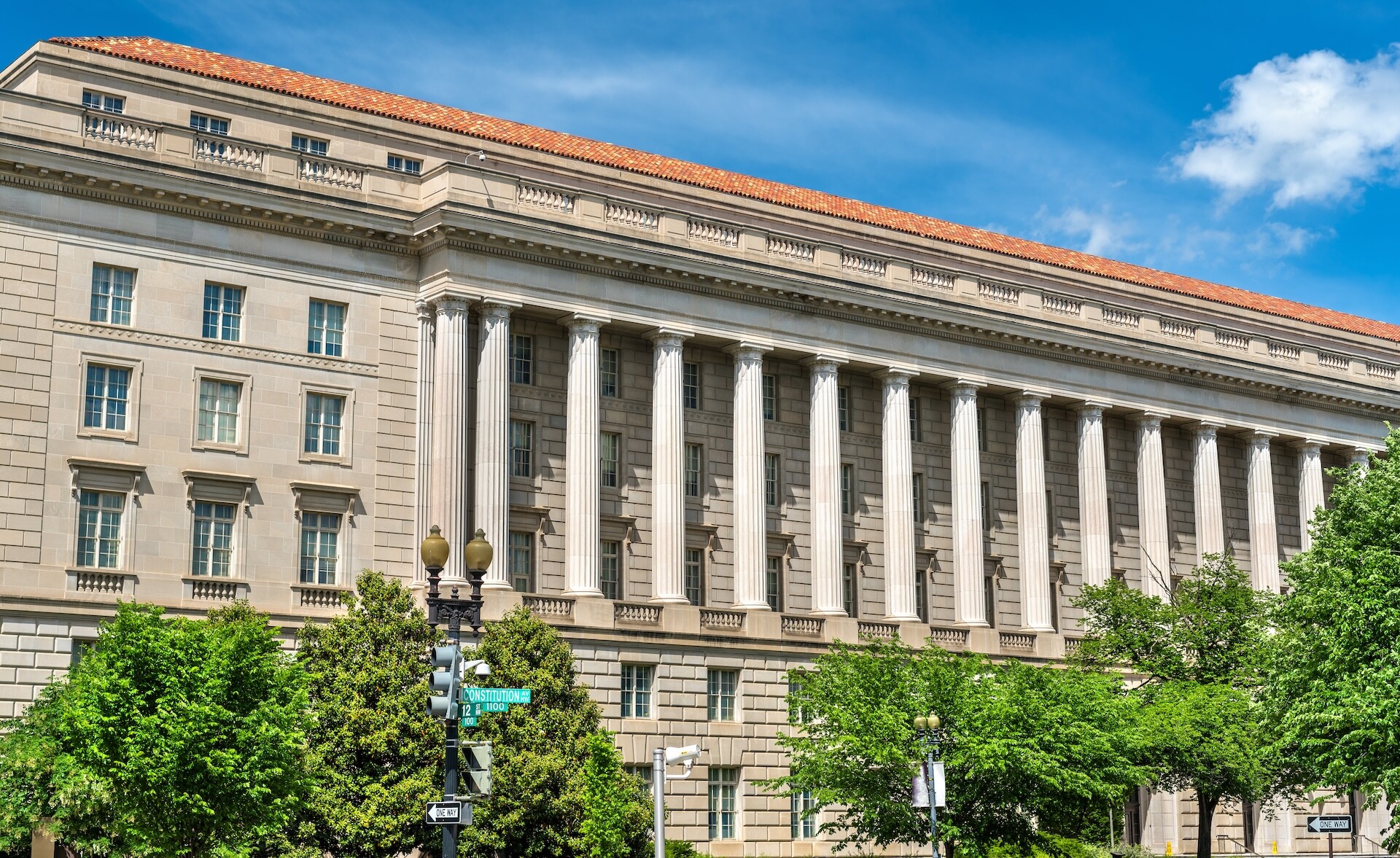 A large, classical-style stone building with tall columns spans the street. It's surrounded by green trees under a clear blue sky with a few clouds. Street lamps and signs are visible in the foreground.