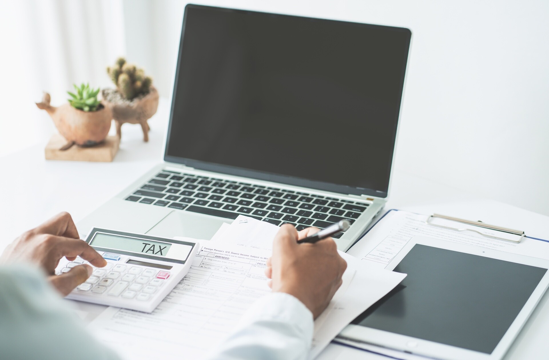 A person is using a calculator displaying the word "TAX" alongside a pen and documents on a desk. A laptop with a blank screen and a tablet on a clipboard are in the background. A small plant decorates the workspace.