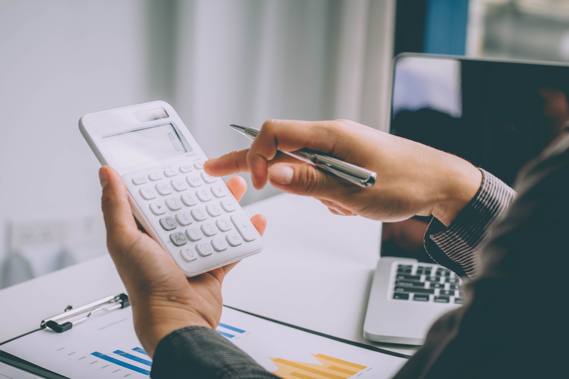 A person holding a white calculator in one hand and a silver pen in the other, sitting at a desk with documents and a laptop. A graph is visible on the documents, suggesting a financial or analytical setting.