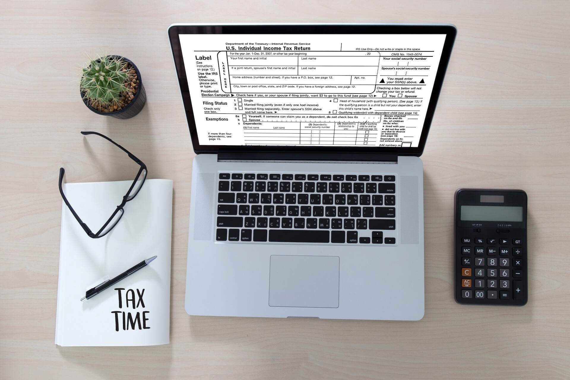 A laptop displaying a U.S. tax form sits on a desk alongside a calculator, a notebook labeled "TAX TIME," and a pair of glasses. A small potted cactus is also on the desk.