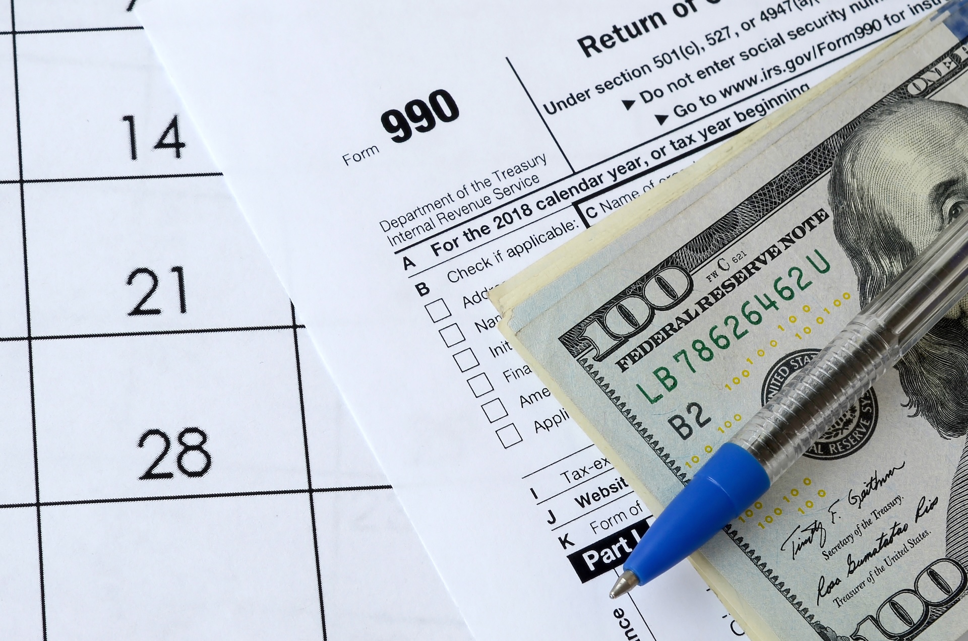 A pen rests on a stack of U.S. 100 dollar bills placed over a Form 990 document. The edge of a calendar page showing the numbers 14, 21, and 28 is visible in the background.