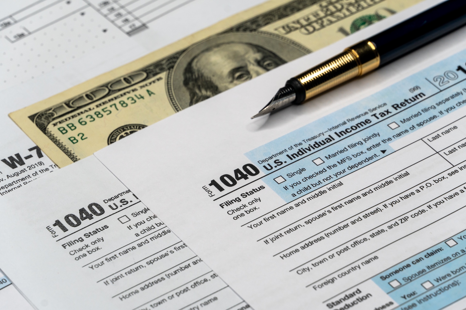 A pen rests on top of a stack of tax forms, including a 1040 form, with U.S. currency visible underneath. The scene suggests financial planning or tax preparation.