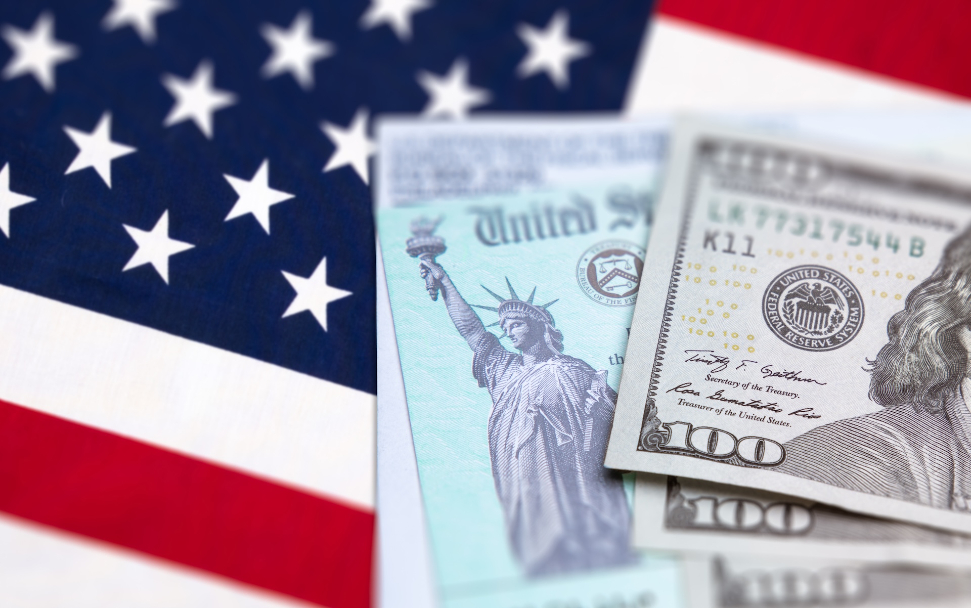 Close-up of a U.S. flag in the background, with a government-issued check and a hundred-dollar bill in the foreground. The Statue of Liberty is visible on the check, symbolizing financial aid or payment.