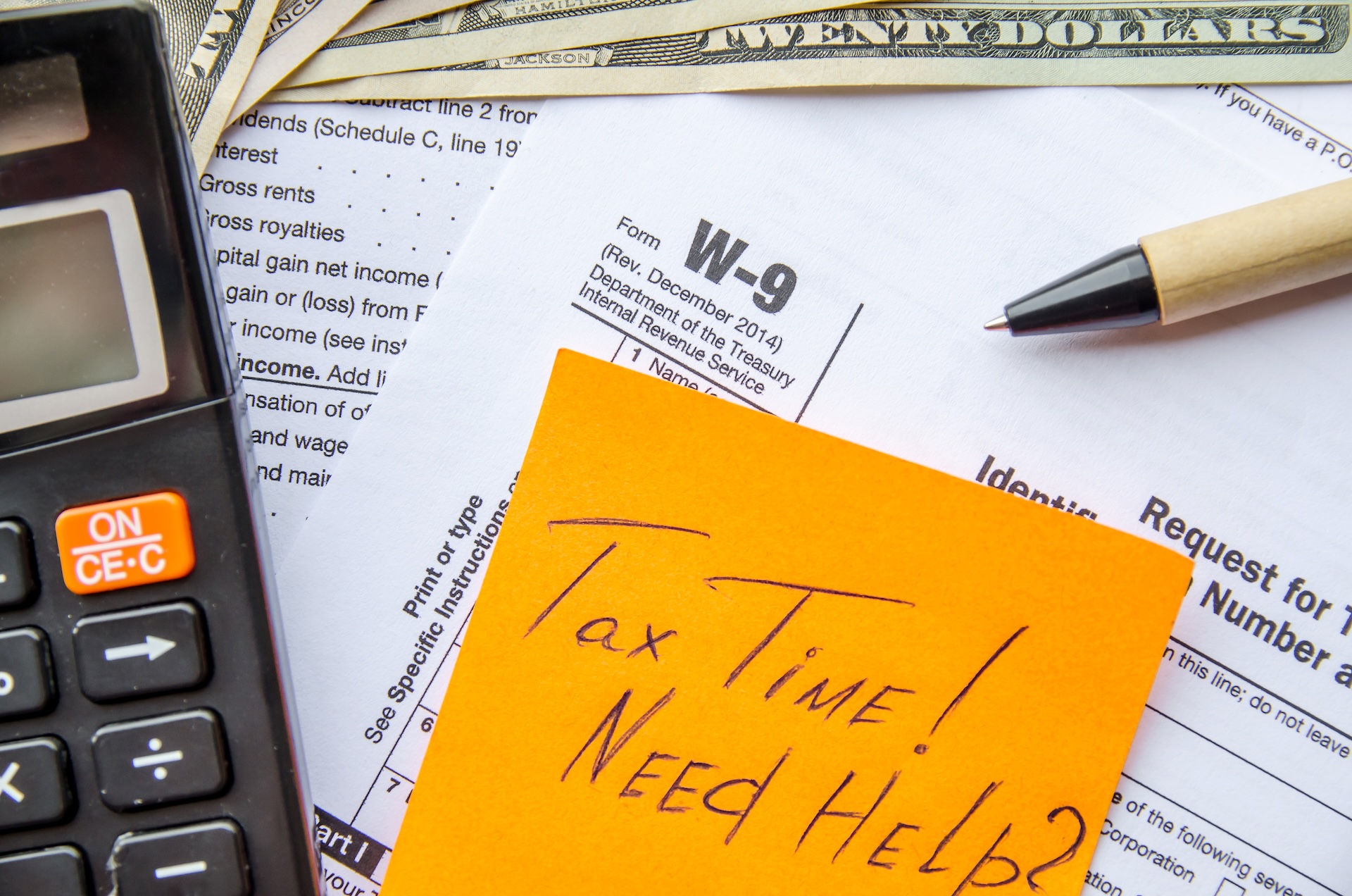 A calculator, pen, and various U.S. tax documents, including a W-9 form, are spread out on a table. A yellow sticky note on top reads "Tax Time! Need Help?" with some U.S. twenty-dollar bills nearby.