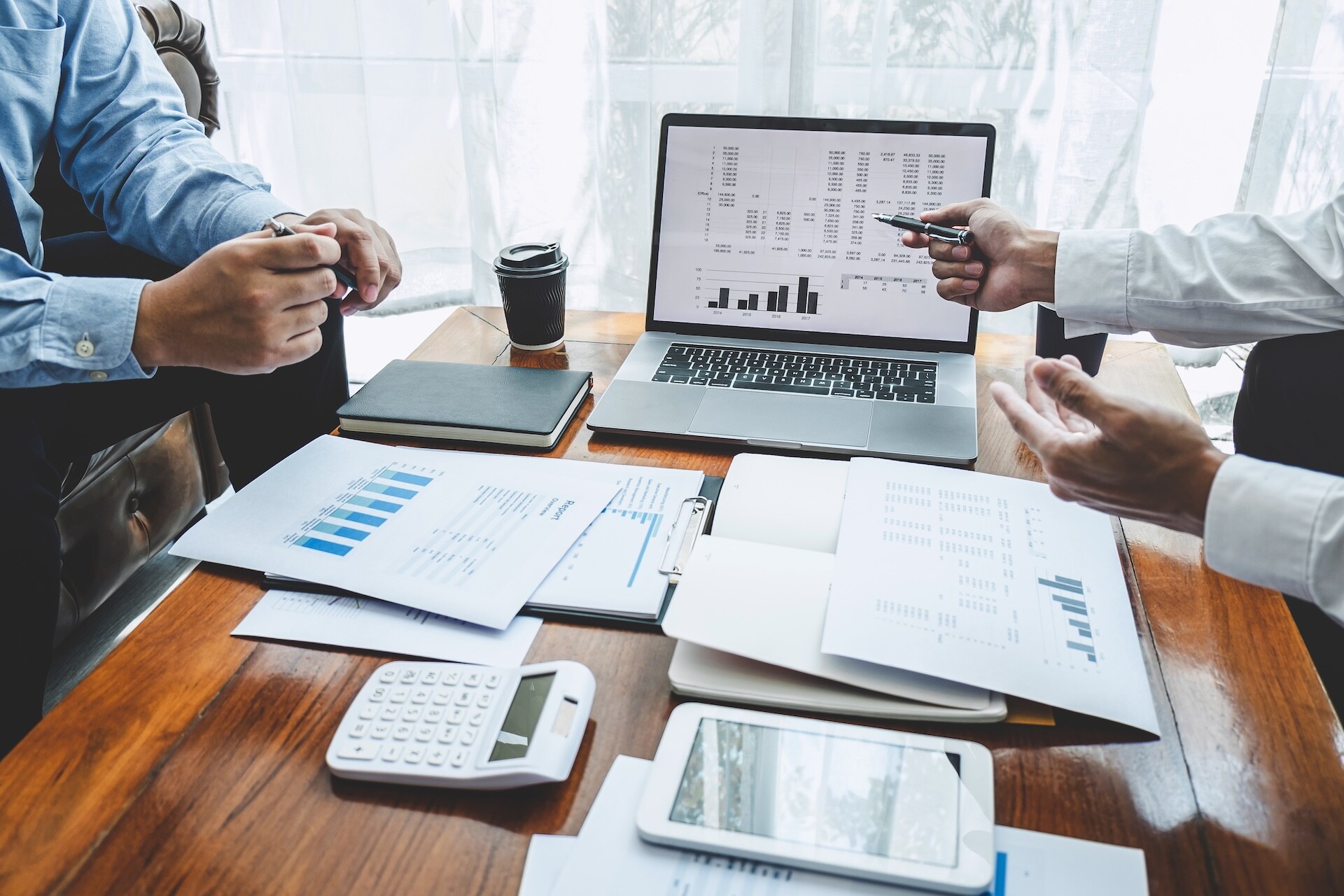 Two people sitting at a table analyzing financial charts and graphs on papers and a laptop. A calculator and a tablet are also on the table alongside a notebook and a coffee cup. One person points at the laptop screen.