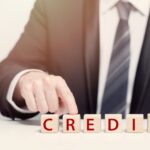 A person in a suit points to wooden blocks on a table that spell "CREDIT" in red capital letters, visually highlighting the concept of What is A Business Tax Deduction vs. A Tax Credit. The background is neutral and out of focus.