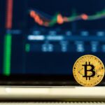 A gold Bitcoin coin stands upright on a surface in front of a laptop displaying a blurry cryptocurrency price chart and market data—highlighting the complexities that may require a business tax debt lawyer in 2025.