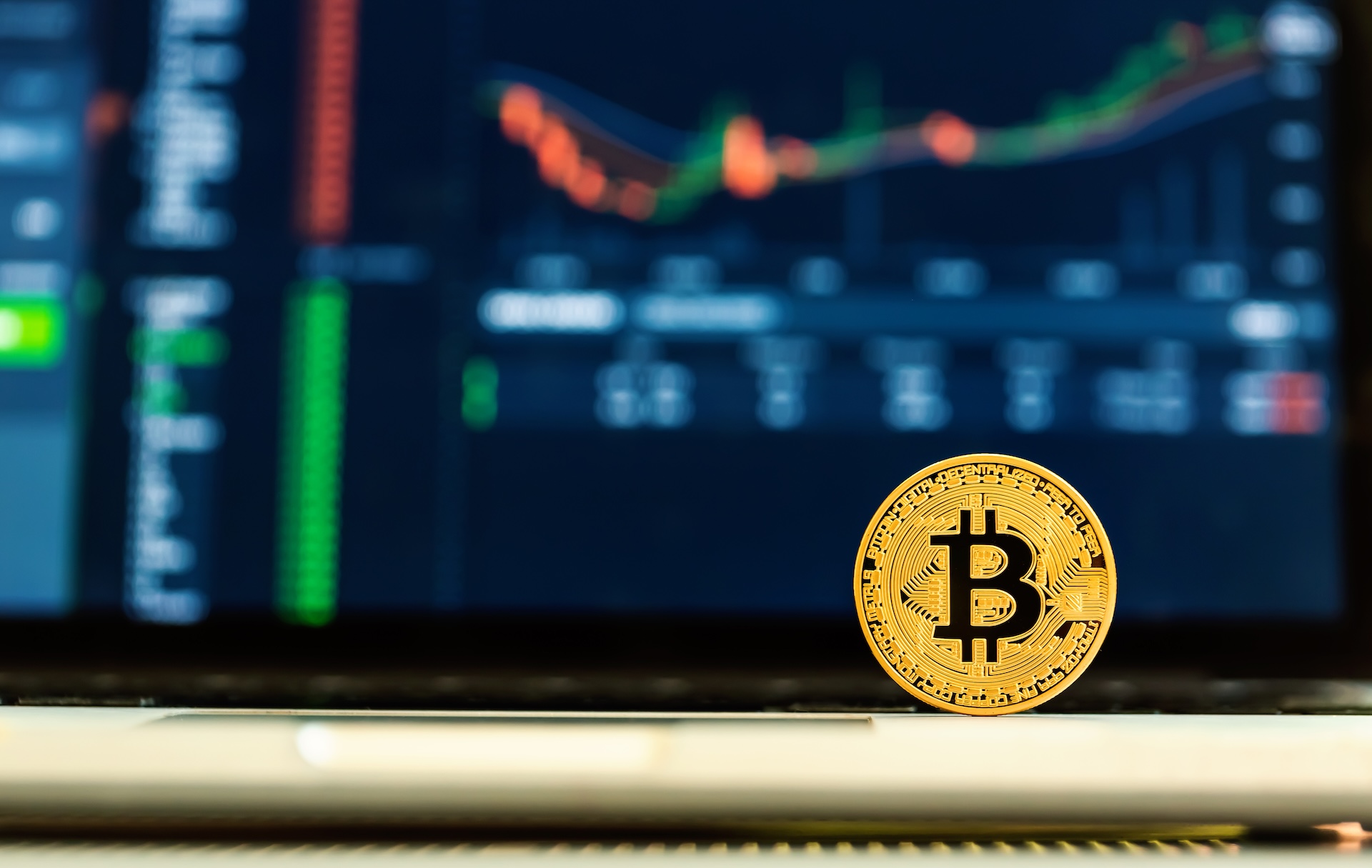A gold Bitcoin coin stands upright on a surface in front of a laptop displaying a blurry cryptocurrency price chart and market data—highlighting the complexities that may require a business tax debt lawyer in 2025.
