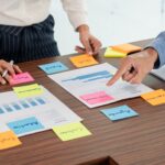 Two people reviewing documents, charts, and colorful sticky notes labeled with tasks like “Do I Need to Collect Sales Tax? Everything Business Owners Should Know,” as they plan and discuss data, charts, and business strategies at a wooden table.