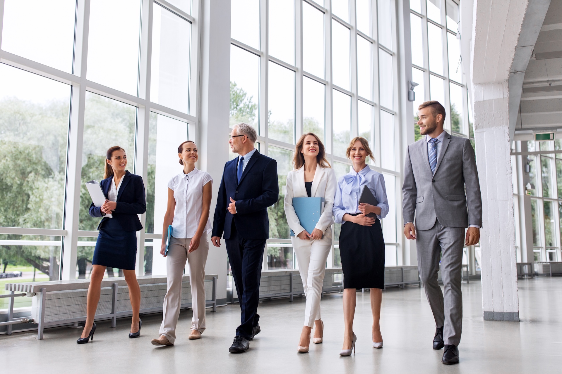 Six business professionals, three women and three men, walk together in a bright, modern office hallway with large windows. They are dressed in formal attire and appear to be engaged in conversation. Six business professionals, three women and three men, walk together in a bright, modern office hallway with large windows. They are dressed in formal attire and appear to be engaged in conversation.