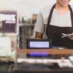 A smiling barista wearing a brown apron stands behind a counter, holding receipts—perhaps pondering what is a business tax deduction vs. a tax credit. A digital cash register screen and a tip box labeled "TIP BOX" are visible in the foreground.