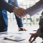 Two people in business attire shaking hands over a table with documents and charts, highlighting a successful business agreement. This scene reflects the importance of strategic planning, like the 10 Reasons Why Every Business Should Have a Tax Plan.