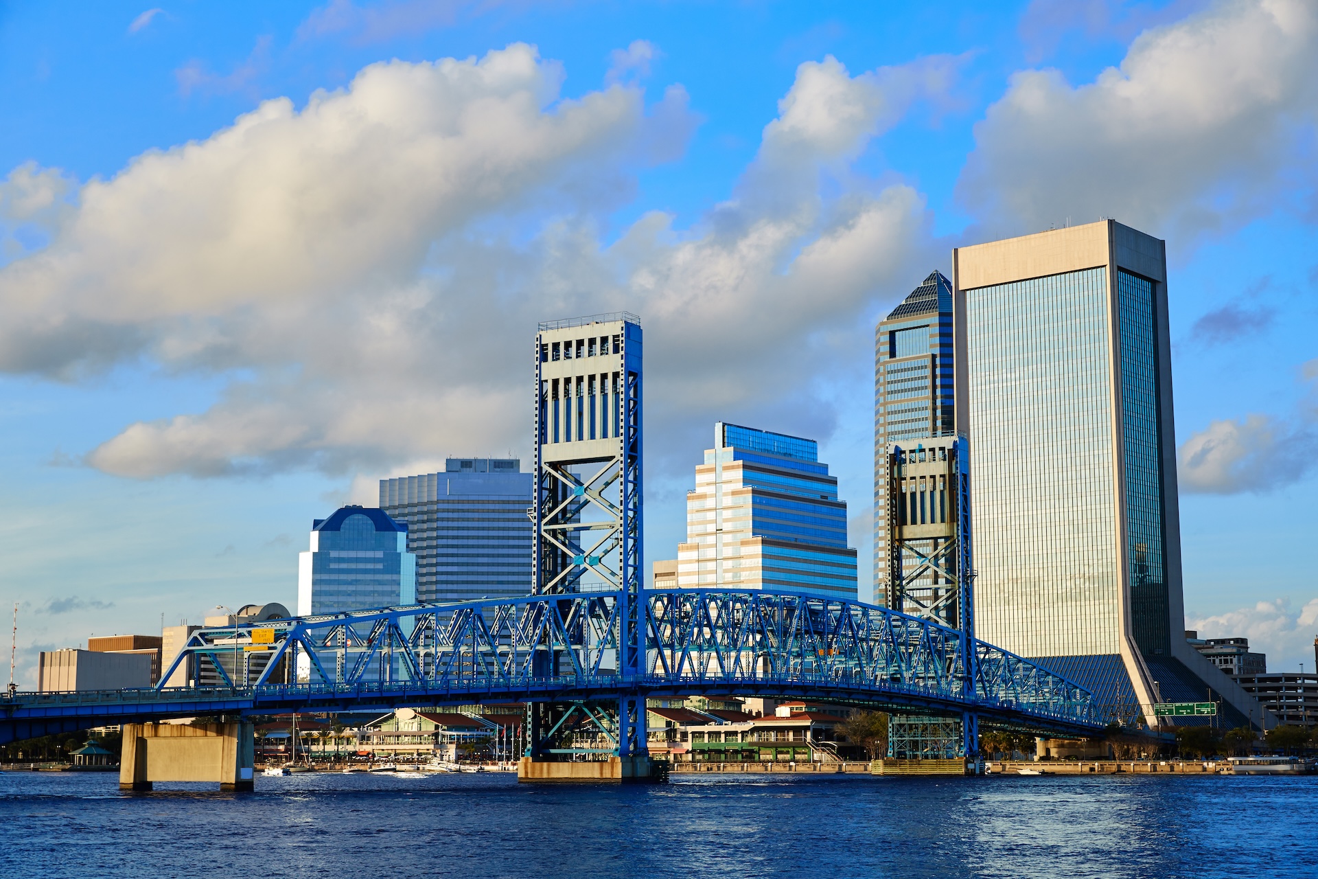 A blue steel bridge spans a river in front of modern high-rise buildings and skyscrapers under a partly cloudy sky.