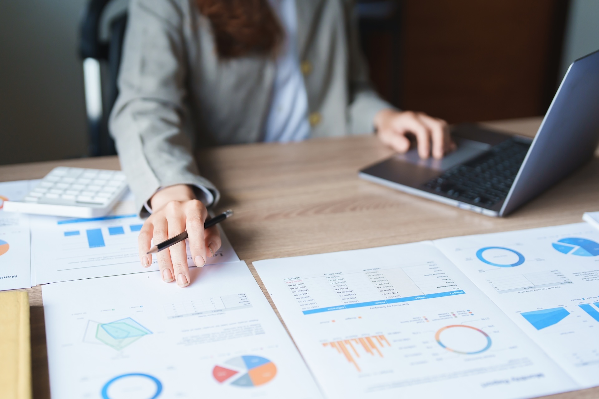 A person in business attire sits at a desk with charts and graphs, pointing at a document with a pen and using a laptop. Financial and statistical reports are spread out on the table. A person in business attire sits at a desk with charts and graphs, pointing at a document with a pen and using a laptop. Financial and statistical reports are spread out on the table.