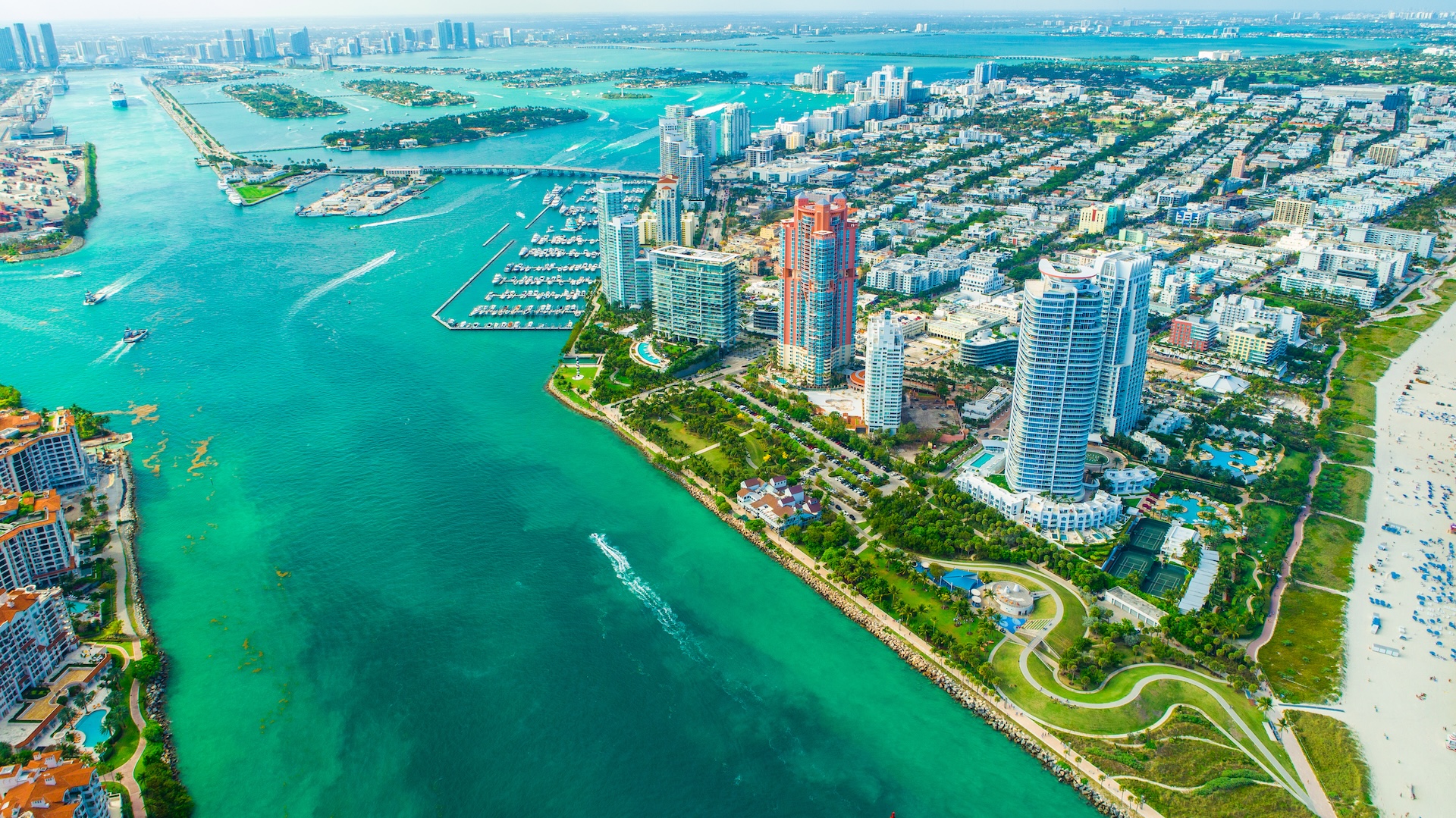 Aerial view of Miami Beach, featuring turquoise water, boats, high-rise buildings, green parks, and sandy beaches along the coastline on a bright sunny day. Aerial view of Miami Beach, featuring turquoise water, boats, high-rise buildings, green parks, and sandy beaches along the coastline on a bright sunny day.