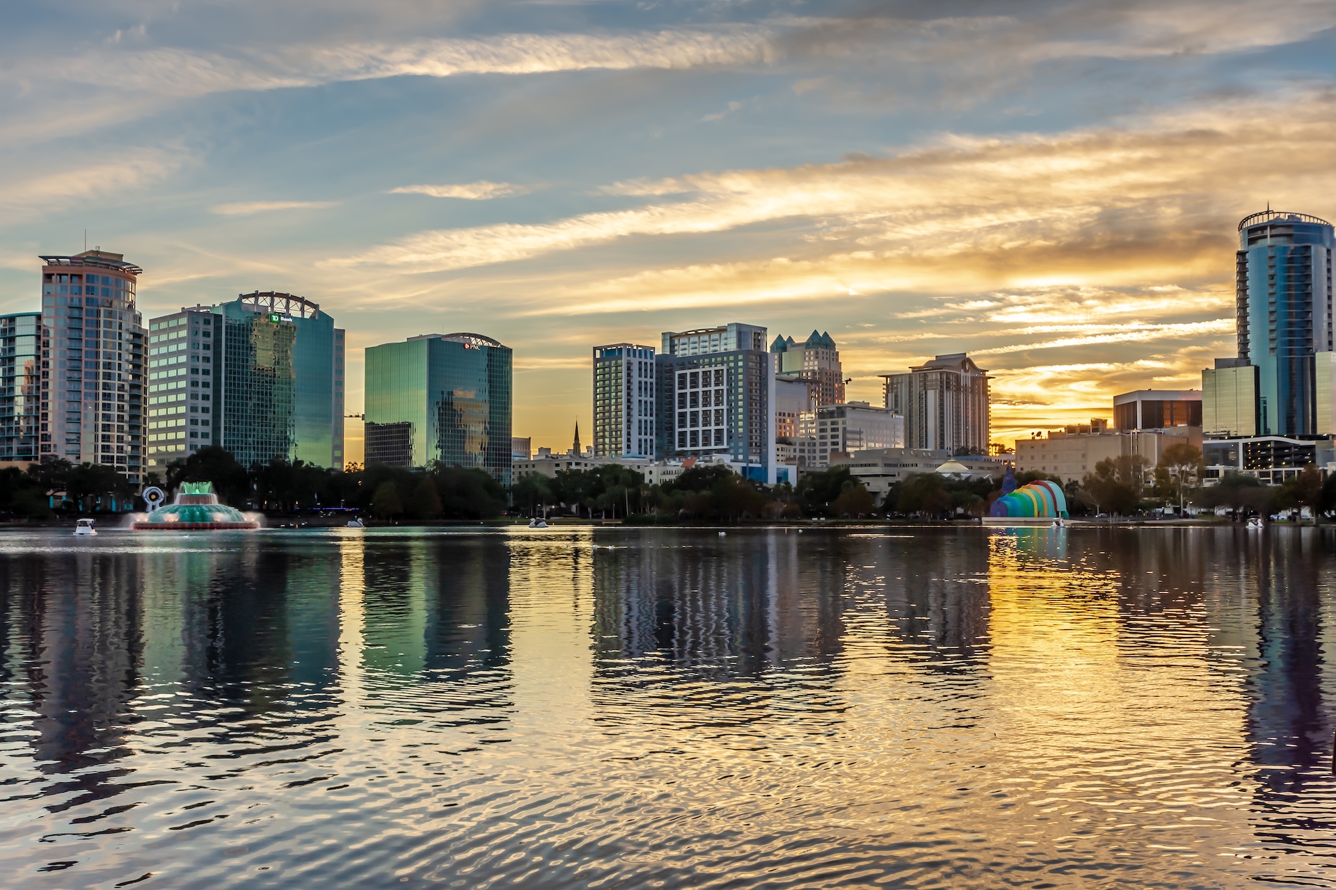 Skyline of a city with modern high-rise buildings reflected in a calm lake at sunset, with clouds in the sky and a fountain and colorful dome visible near the water’s edge.