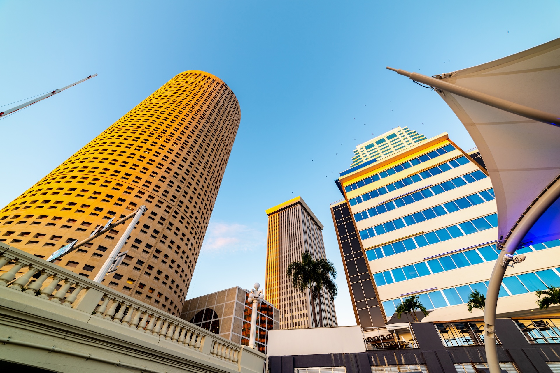 Modern skyscrapers and office buildings rise against a clear blue sky, with palm trees and architectural details in the foreground, creating a vibrant cityscape at sunset.
