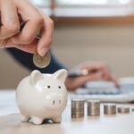 A hand places a coin into a white piggy bank, with stacked coins and a person using a calculator in the background, symbolizing saving money, financial planning, and understanding IRS notices and how to respond.