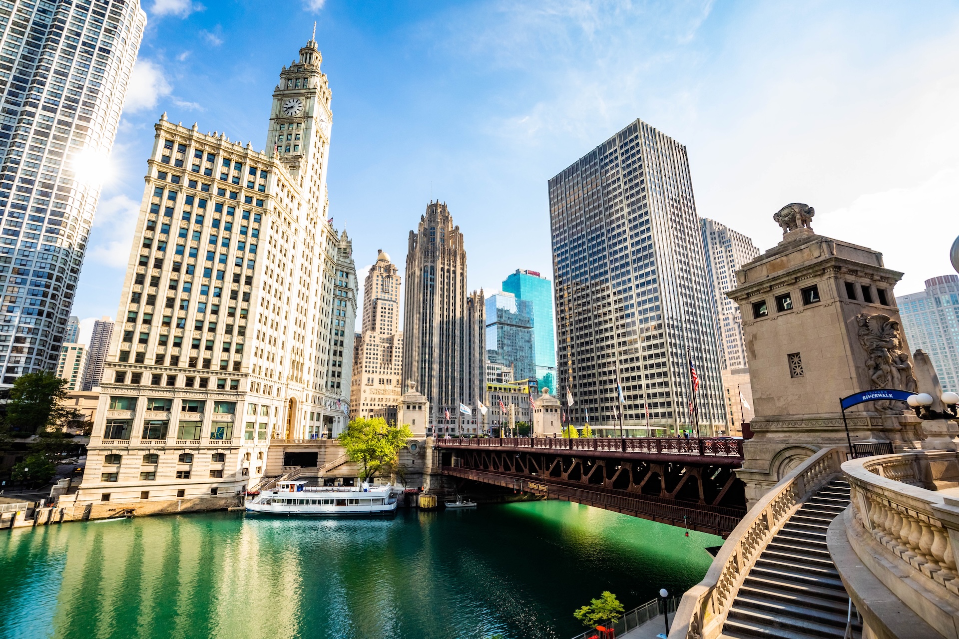 Chicago Riverwalk view featuring the Wrigley Building, Tribune Tower, high-rise buildings, a boat on the river, and the DuSable Bridge under a sunny sky.