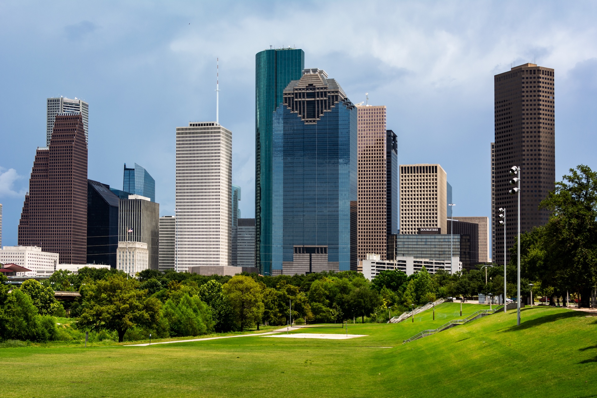 Houston skyline with tall modern skyscrapers and glass buildings, seen from a green park with trees and a grassy open area under a cloudy blue sky.