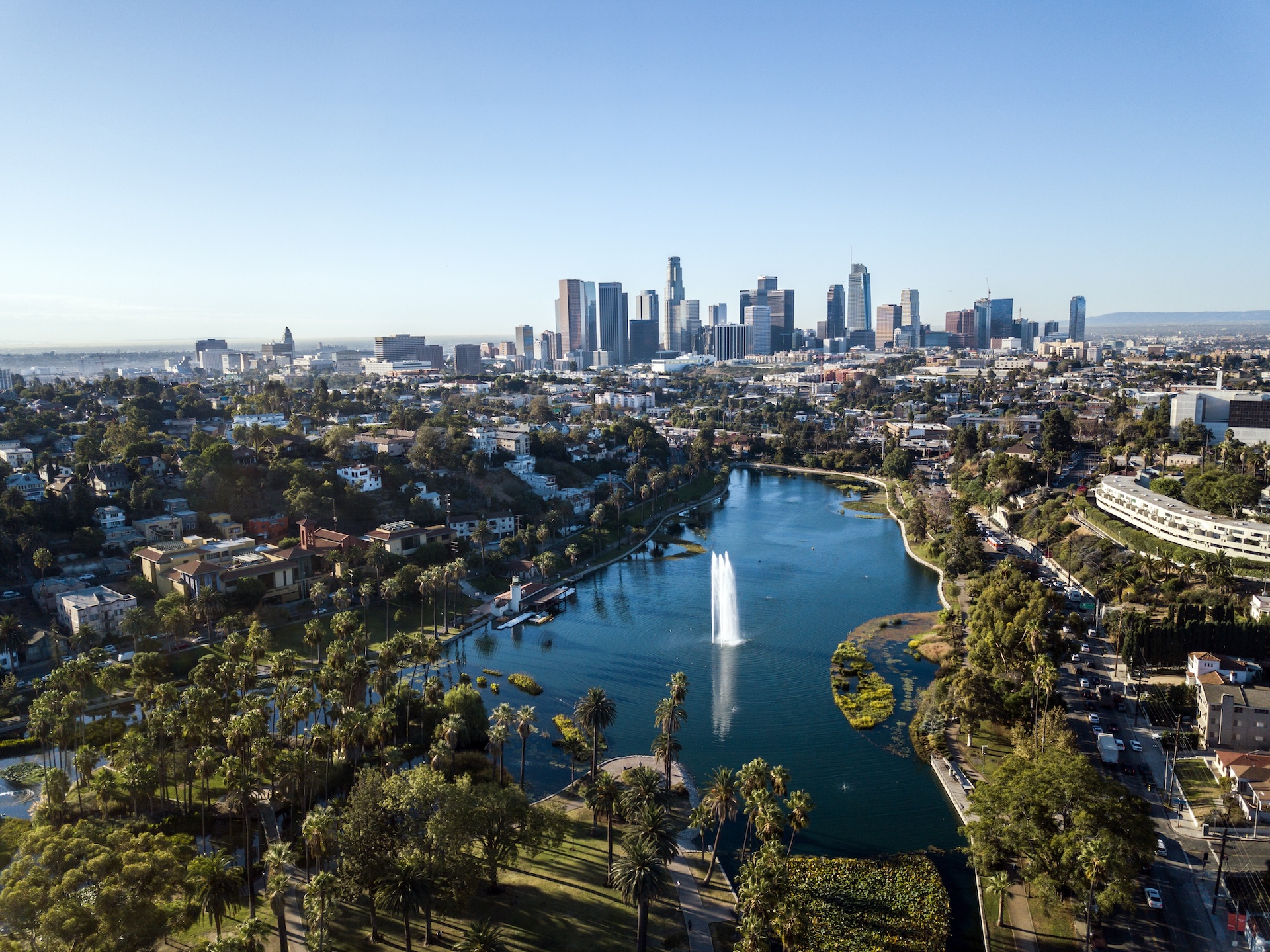 Aerial view of downtown Los Angeles skyline with tall skyscrapers, Echo Park Lake with fountains, and palm trees in the foreground on a clear, sunny day.
