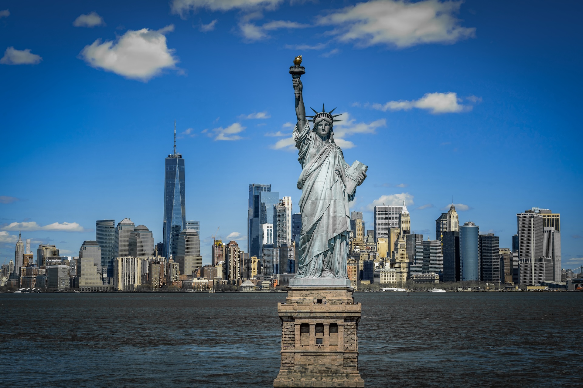 The Statue of Liberty stands on Liberty Island with the Manhattan skyline, including One World Trade Center, visible across the water under a blue sky with scattered clouds.