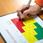 A hand holding a pen fills out a risk assessment chart on a desk, highlighting colored sections for different risk levels—a visual reminder of when should a business consider a risk assessment to safeguard operations.