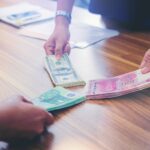 Three people exchange bundles of US dollars, Chinese yuan, and euros over a wooden table. Only their hands are visible, while documents—possibly related to FACTA & GILTI Explained—are blurred in the background.