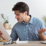 A man sitting at a desk looks confused and frustrated while holding a piece of paper in one hand and gesturing with the other, with a laptop and notebook in front of him.