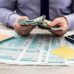 A person in a dress shirt counts cash at a desk filled with tax forms, a laptop, sticky notes, and a calculator—wondering, "Are You Eligible to Settle Tax Debt? Let a Tax Attorney Show You the Way.