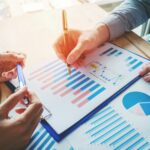 Two people review charts and graphs with blue and red bars and pie charts on paper. One points with a pen, while the other holds a clipboard, highlighting why businesses earning $50K+ need tax planning now.